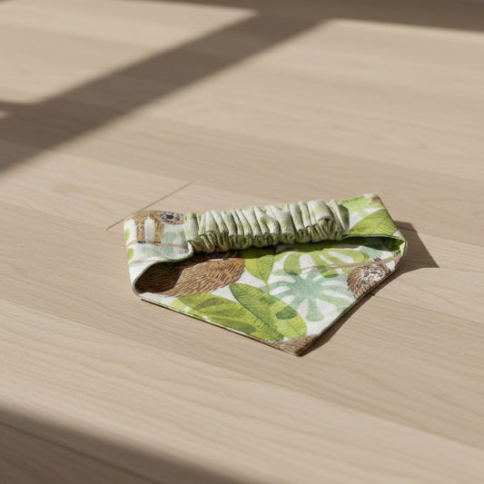A triangular bandana bib with a green jungle print featuring sloths and leaves, and a gathered elastic neck strap, laid flat on a textured off-white background.