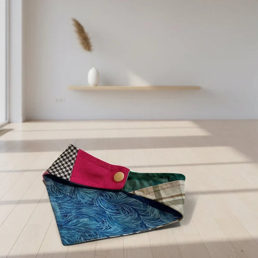 A handmade patchwork fabric clutch bag with a snap closure, resting on a light wooden floor in a minimalist room. The bag features multiple fabric panels including a blue marbled pattern, a pink velvet section, a black-and-white checkered pattern, and a green velvet section.